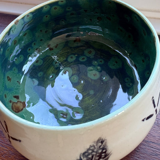 A handmade ceramic bowl with a glossy dark green and teal patterned interior and a cream-colored exterior decorated with black botanical motifs, shown on a wooden surface.
