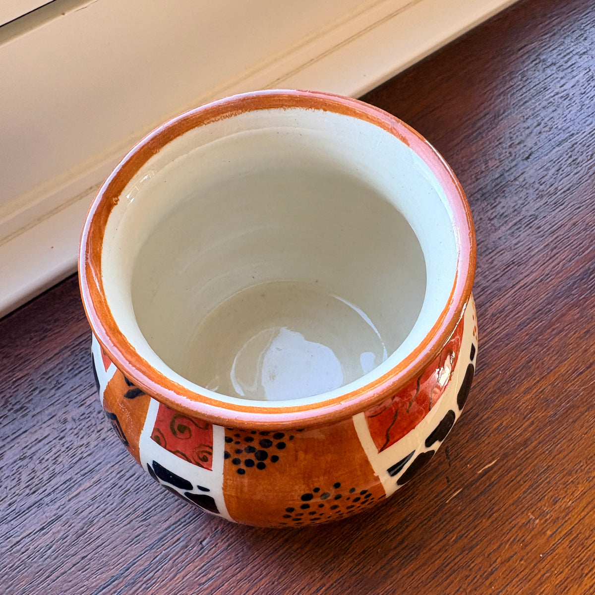 A small handmade ceramic cup with a warm orange-brown exterior patterned with black and red, and a glossy off-white interior, resting on a dark wooden table.