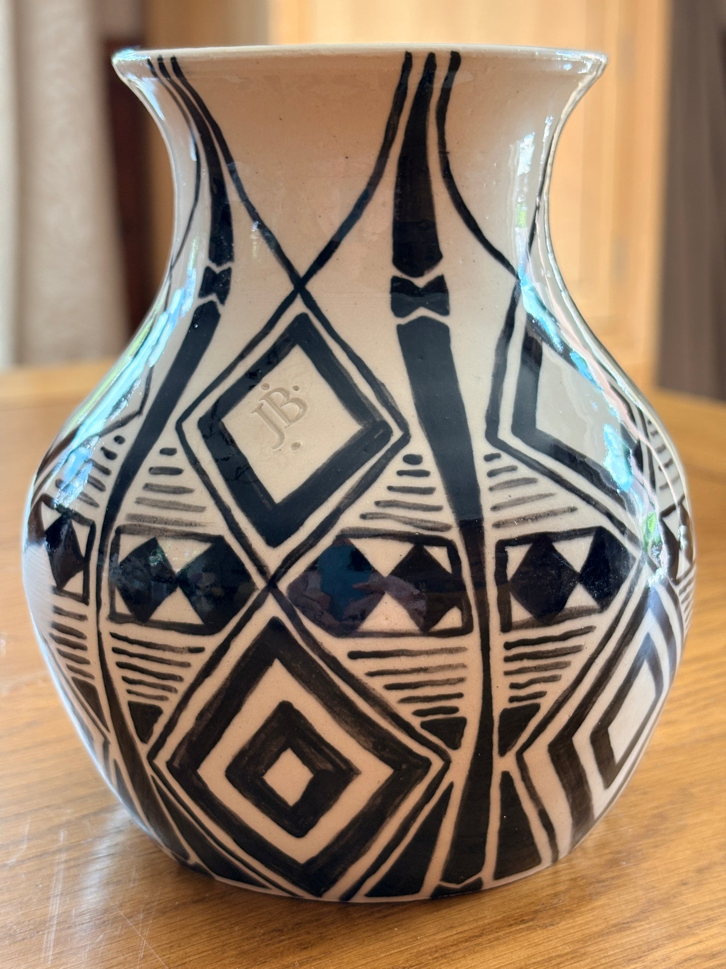 A ceramic vase with a black and off-white geometric pattern, featuring a monogram 'JB' on one of the diamond motifs, resting on a wooden surface.