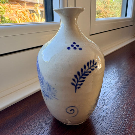 A small ceramic vase with a cream-colored glaze and blue botanical patterns, standing on a dark wooden windowsill.