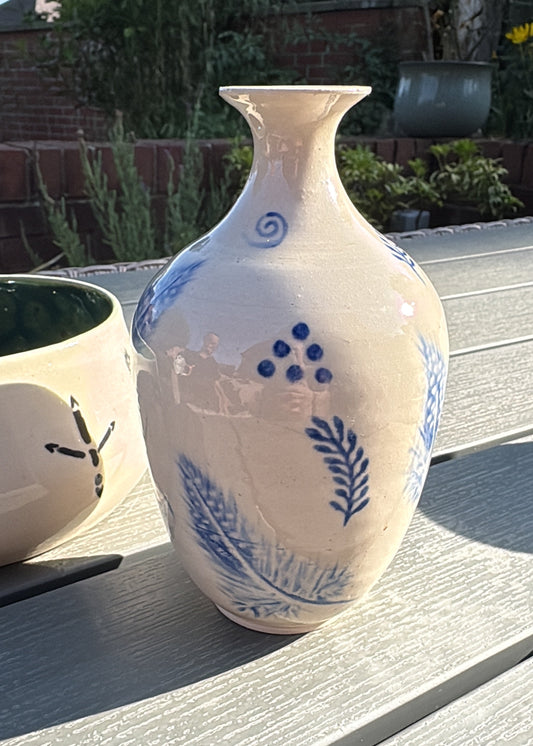 A white ceramic vase with blue painted botanical and grape motifs, sitting on a gray wooden deck outdoors.