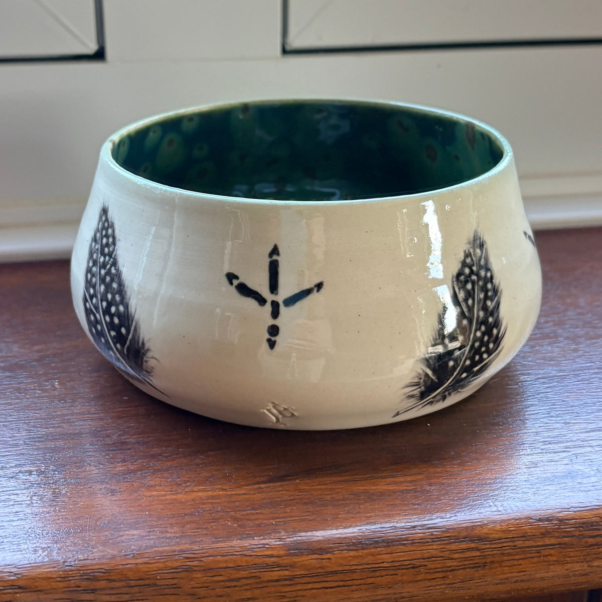 A small ceramic bowl with a cream-colored exterior, black decorative patterns, and a dark green interior, sitting on a wooden windowsill with a window and outdoor view in the background.