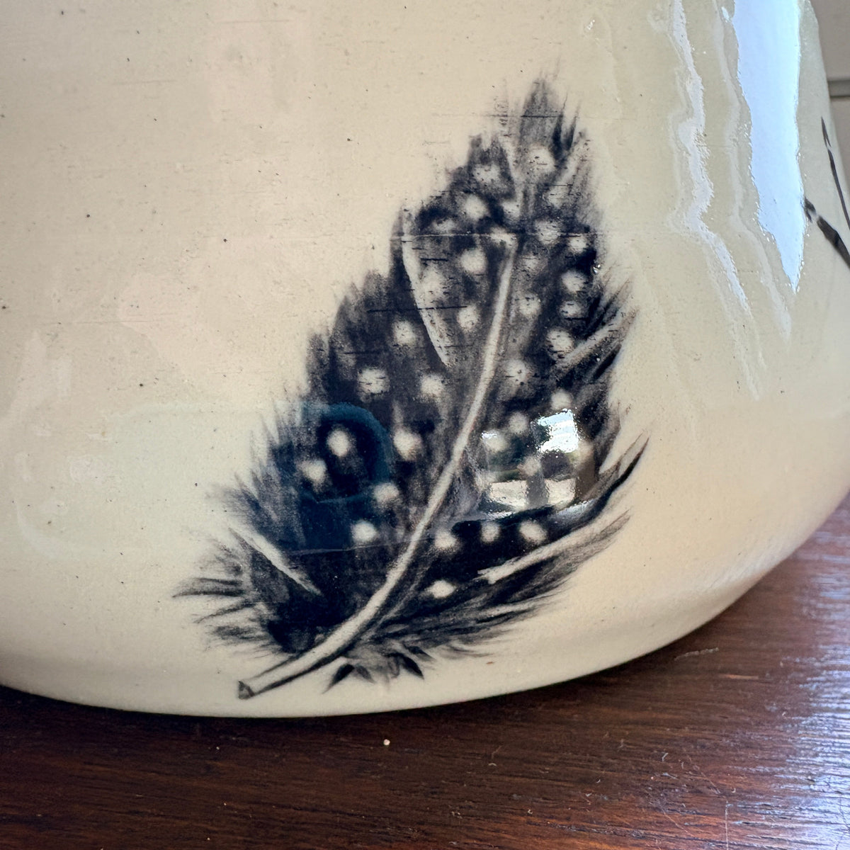 Close-up of a ceramic mug or pot with a black feather design on a cream-colored glaze, resting on a dark wooden surface.