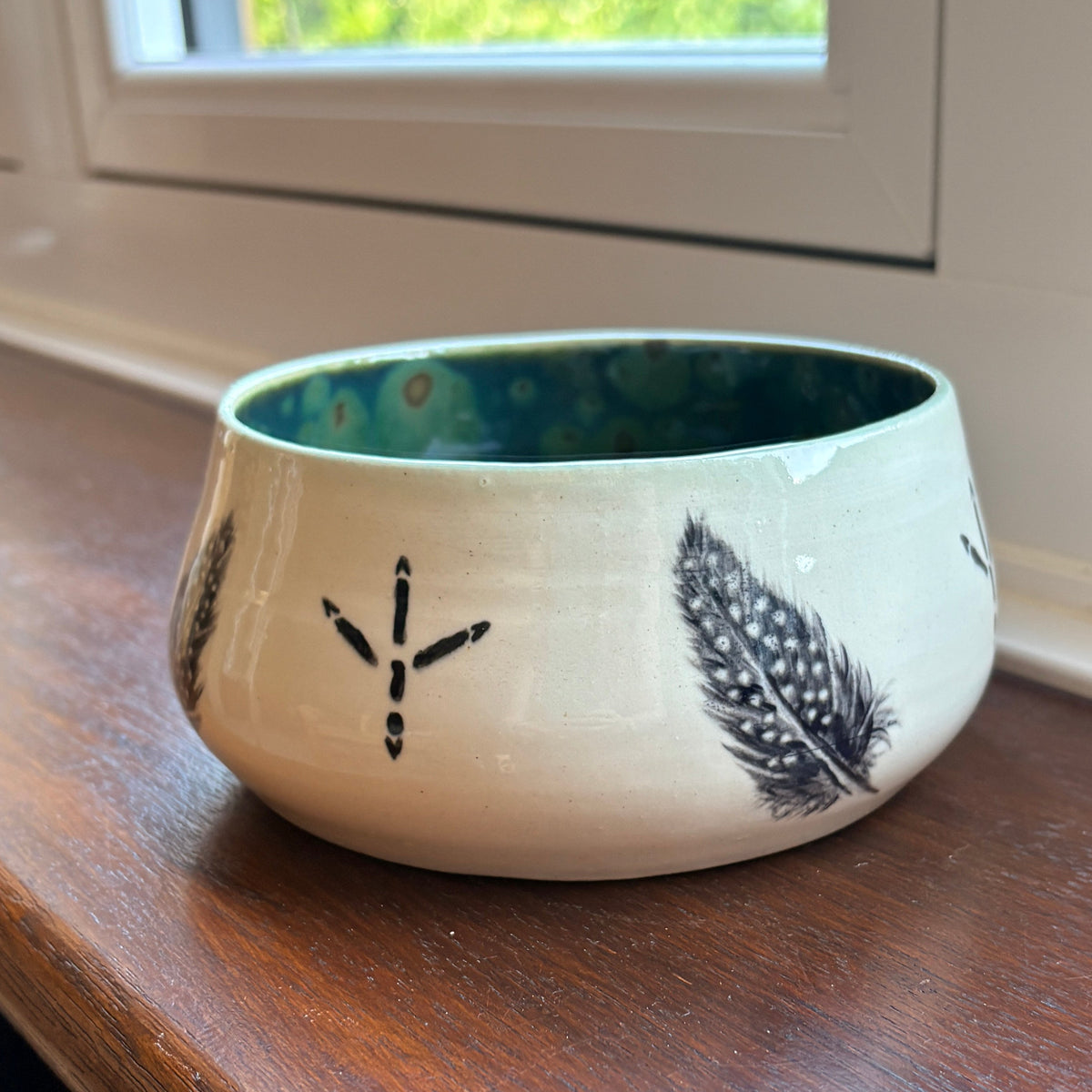 A small ceramic bowl with a cream-colored exterior decorated with black feather and dot patterns, and a dark green glazed interior, sitting on a wooden windowsill.