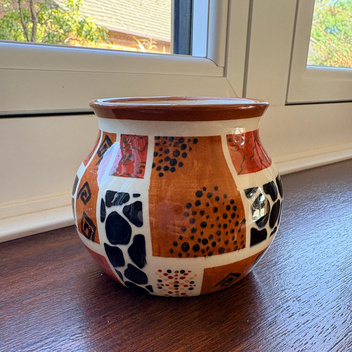 A handmade ceramic pot with a patchwork design of terracotta, black, red, and white patterns, sitting on a dark wooden table near a window.